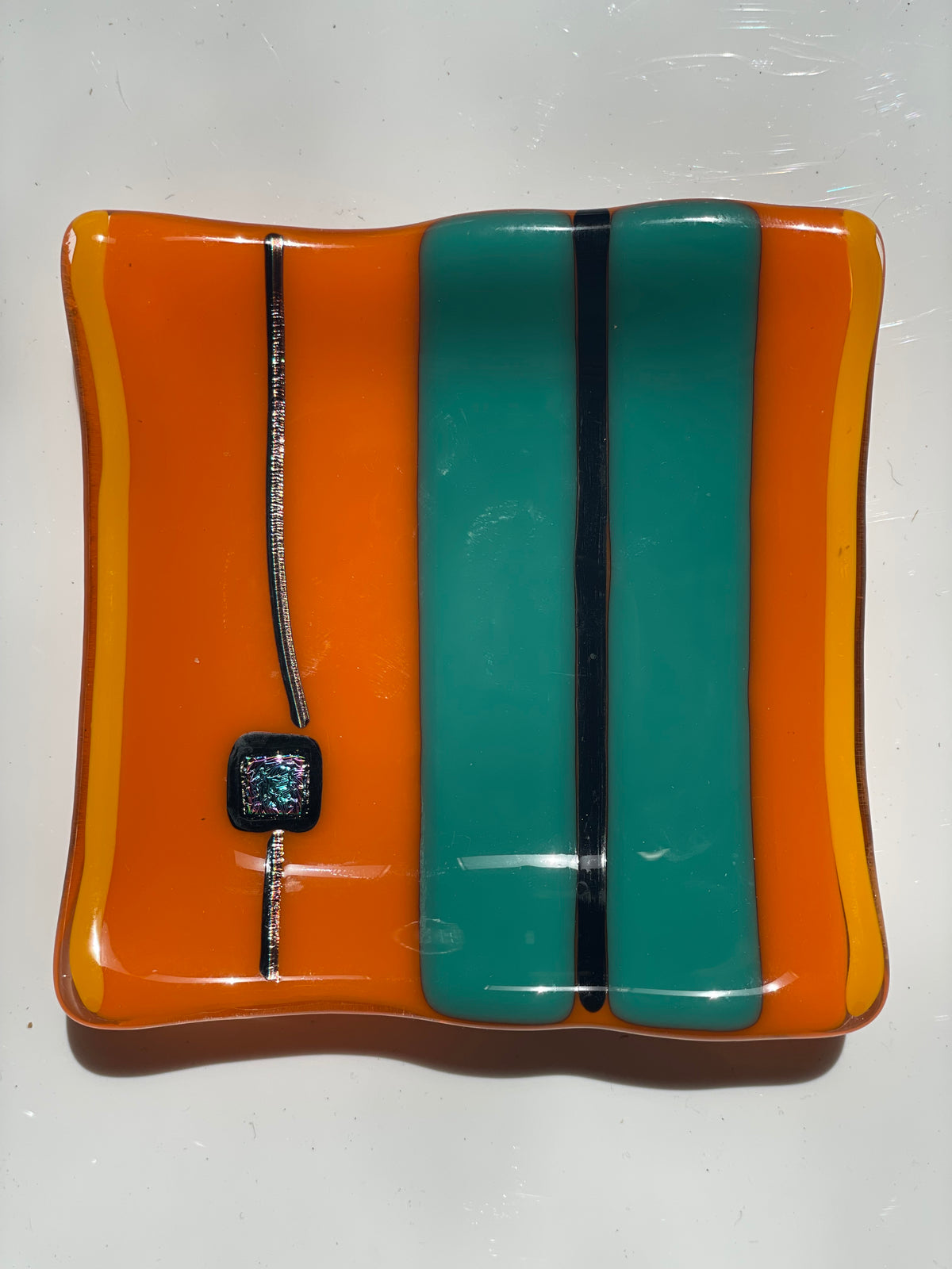 A square fused glass plate or tray with rounded corners, featuring horizontal bands of bright orange, teal green, and yellow, accented by a thin black stripe and a small iridescent square bead on a silver line.