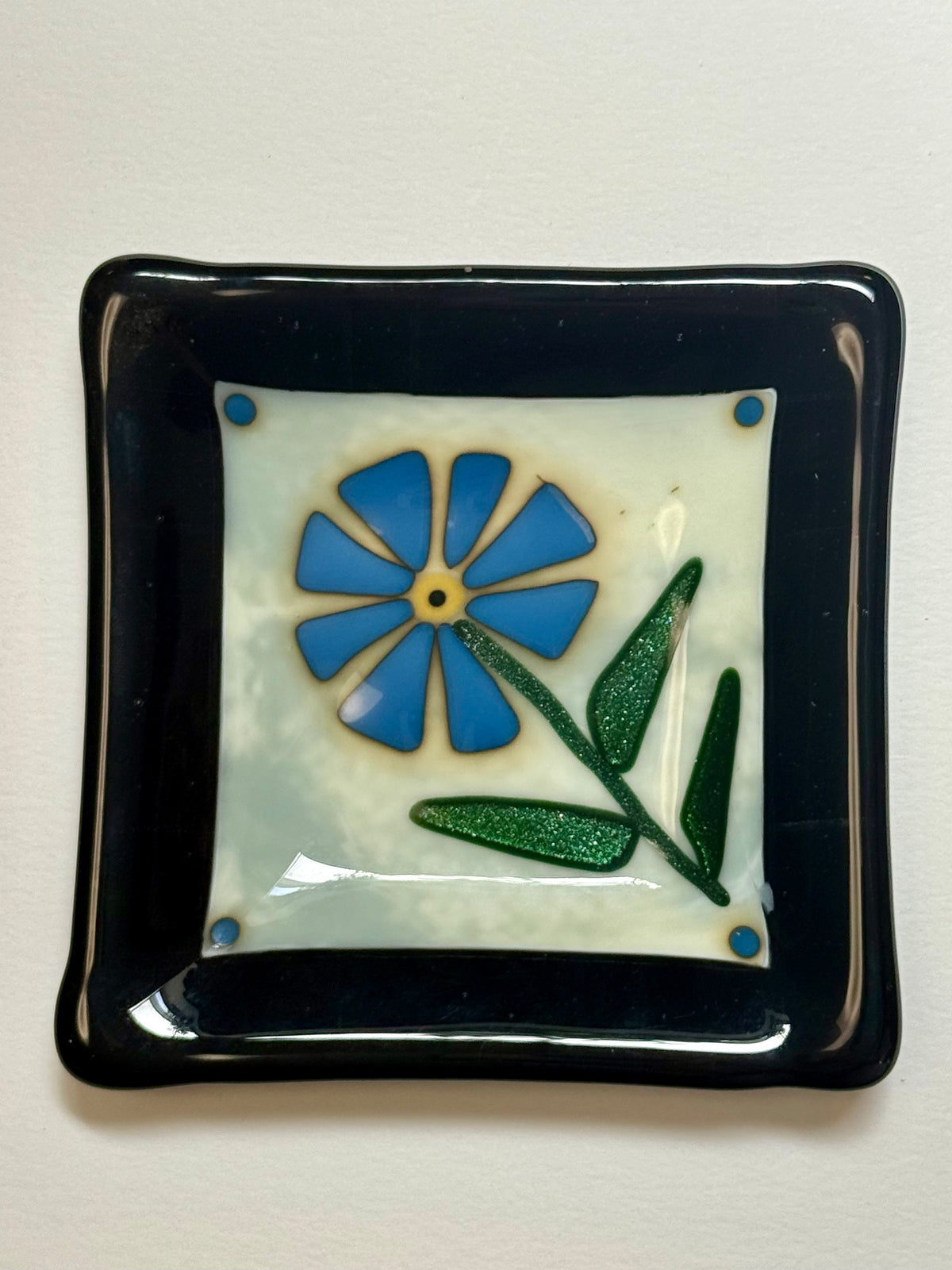 A square glossy fused glass plate with a, off white center panel featuring a blue flower and green leaves.